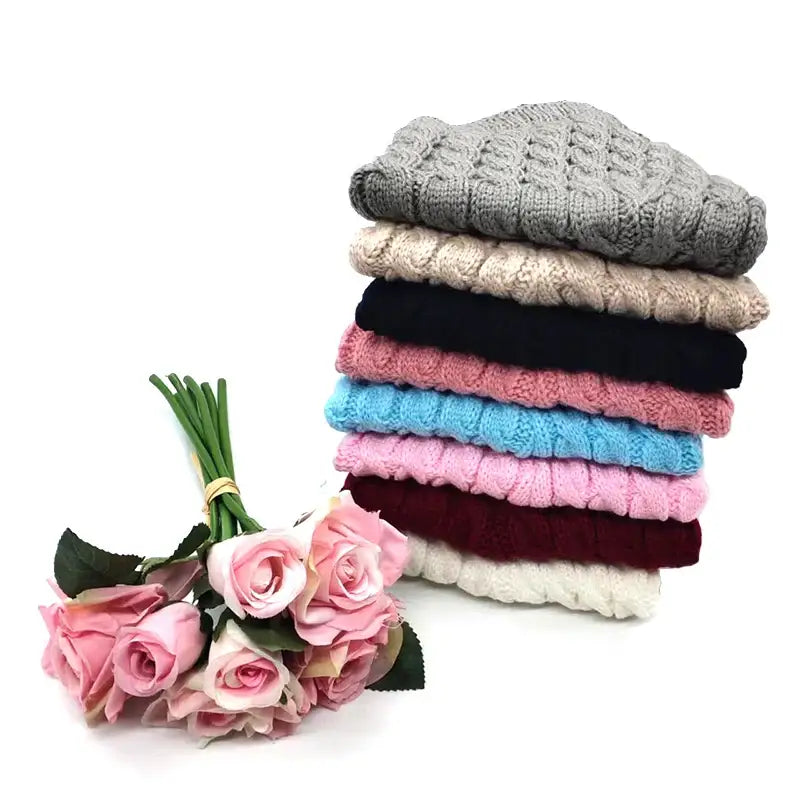 Stack of colorful knitted or crocheted winter hats with a bouquet of pink roses beside them.