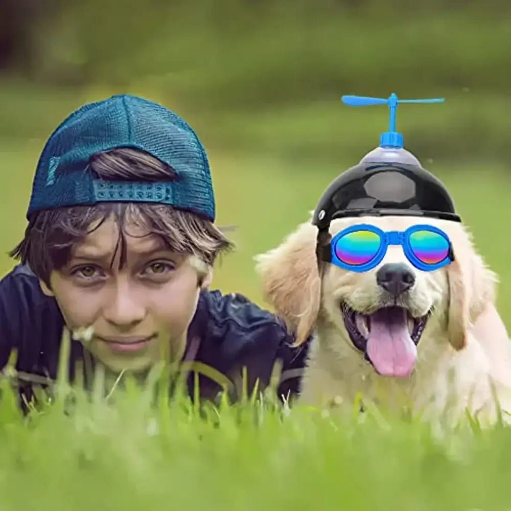 A young person and a dog wearing sunglasses and a propeller hat lying in grass.